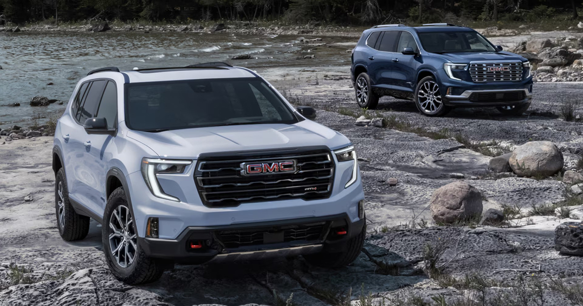Two 2025 GMC Acadia SUVs, white AT4 and blue Denali, positioned on rocky riverbed showcasing capability