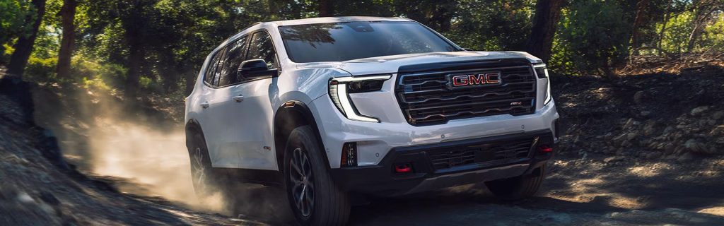 White GMC SUV driving off-road on a dusty forest trail with sunlight filtering through trees.