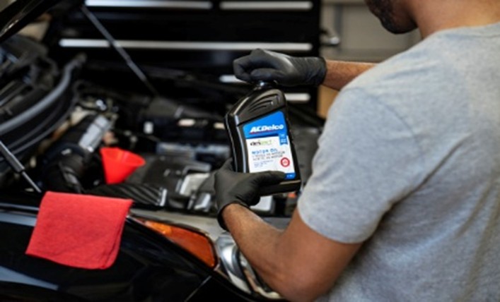 A person wearing black gloves holds a bottle of ACDelco motor oil over an open car engine bay, which features a red funnel and a red shop towel on the fender.