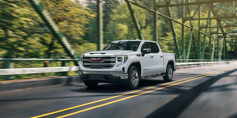 A white GMC Sierra 1500 pickup truck driving across a green steel truss bridge surrounded by lush green trees, captured with a motion-blur effect.
