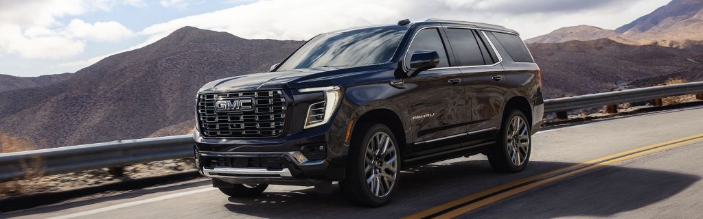 A dark metallic black GMC Yukon Denali SUV driving along a winding mountain road with a deserted mountain landscape in the background.