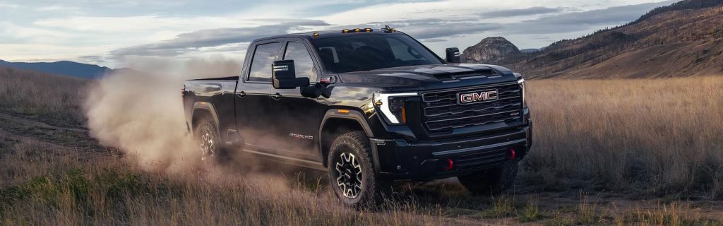 A black GMC Sierra HD AT4X pickup truck driving off-road through a grassy field, kicking up a cloud of dust.